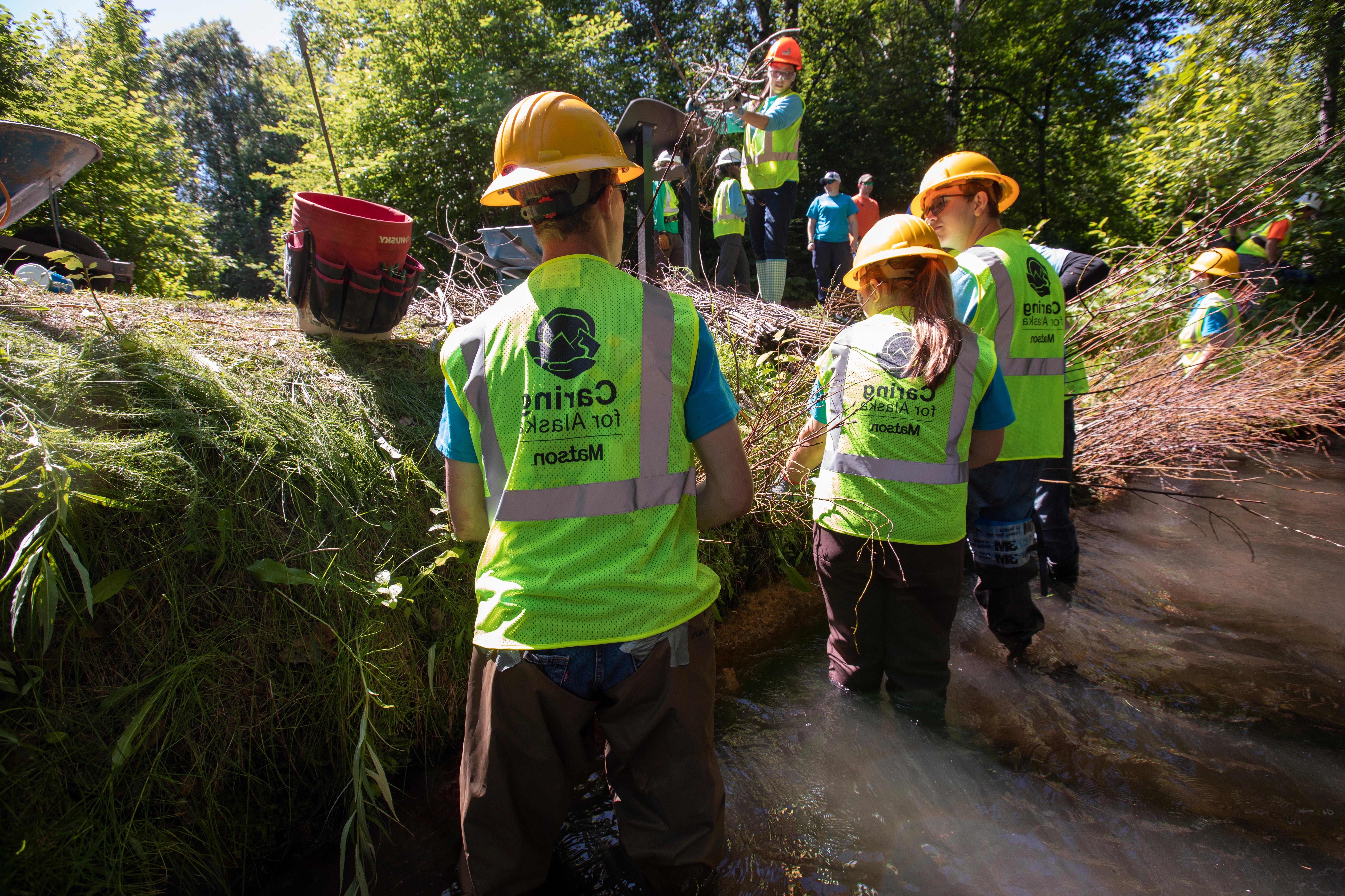 Youth Employment in Parks participants wear green safety vests and yellow hard hats while standing in Chester Creek to remove invasive species and conduct habitat restoration.